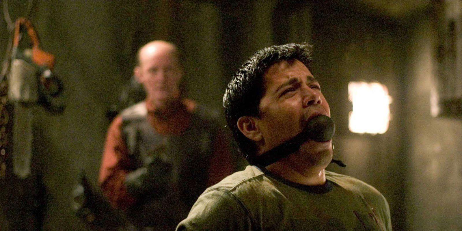 Jay Hernandez with a ball gag in his mouth as an apron-wearing figure stands behind him in Hostel.
