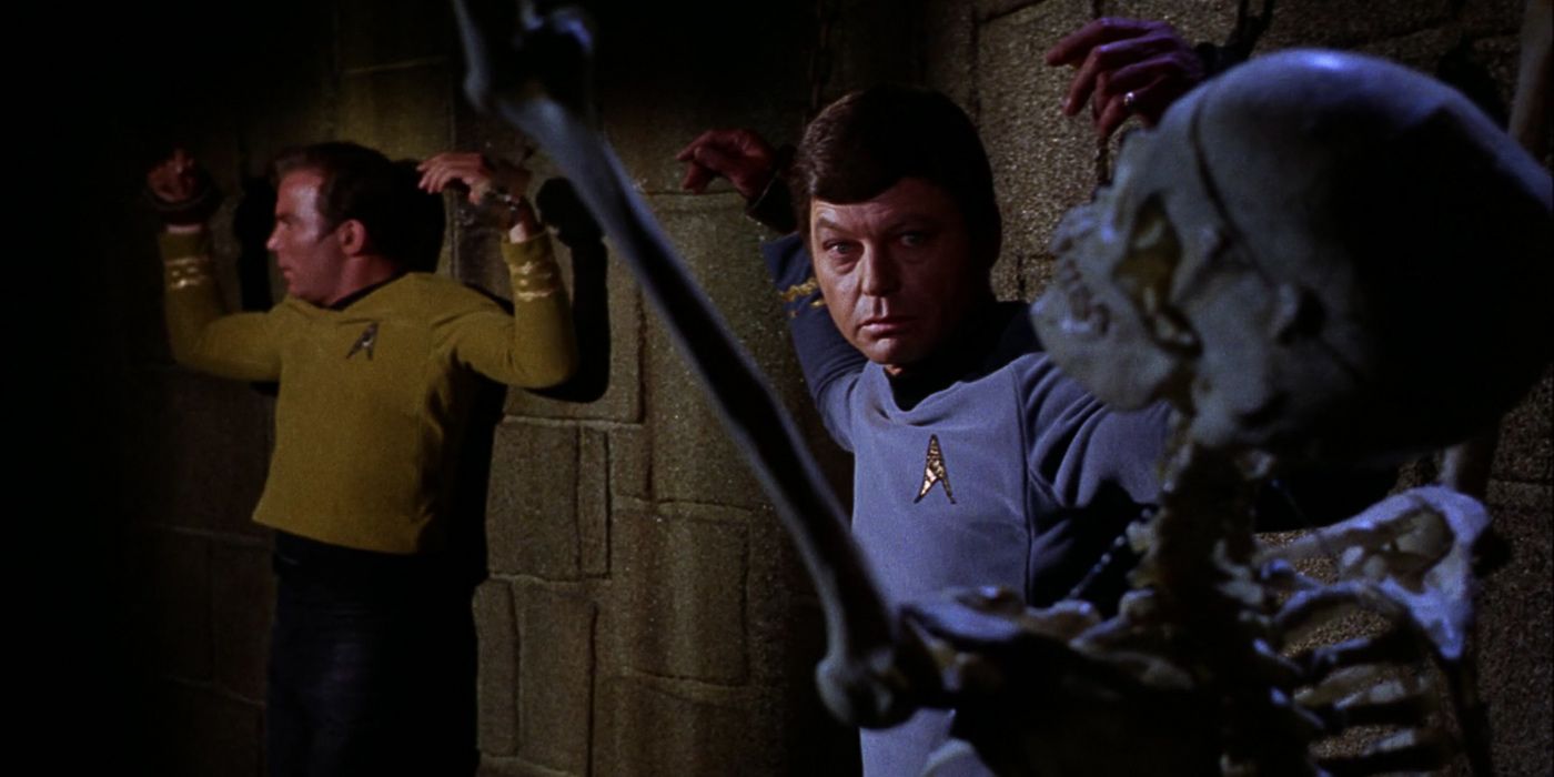Kirk and McCoy stand chained next to a skeleton 