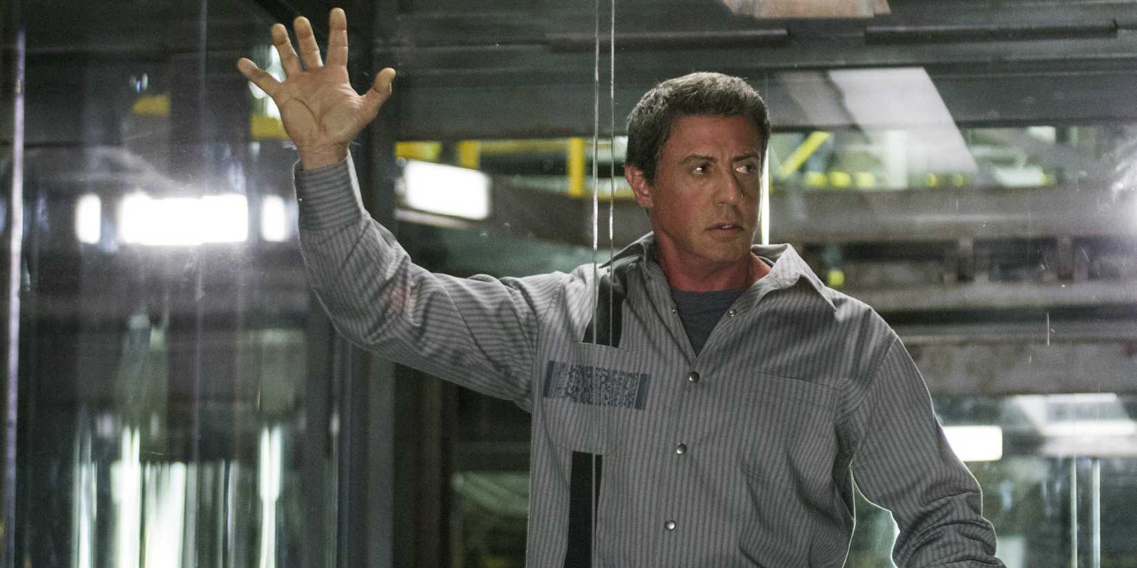 Sylvester Stallone as Ray Breslin holding onto the glass of his cell in Escape Plan