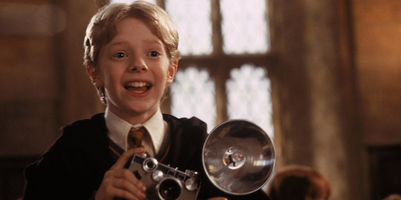 Colin Creevey sorrindo enquanto segura uma câmera no Salão Principal de Hogwarts