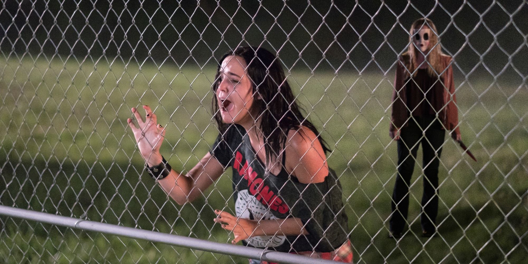 Kinsey standing in front of a fence with a killer behind her in Strangers: Prey At Night