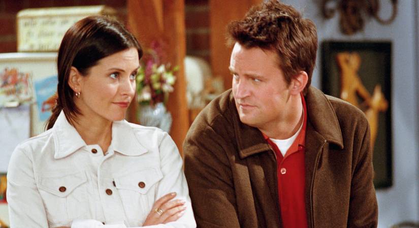 Monica (Courteney Cox) and Chandler (Matthew Perry) of Friends standing side-by-side in their apartment. Chandler looks at Monica while she looks off with her arms crossed.