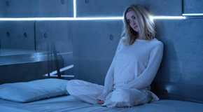 Brianne Howey as Shauna Babcock sitting in a dull gray room in a full white outfit in The Passage