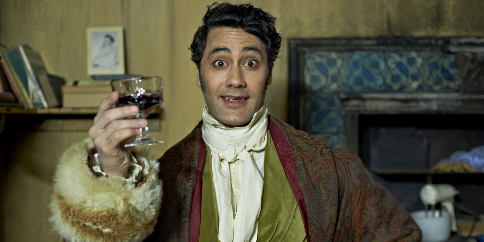 Taika Waititi with vampire teeth holding up a glass in What We Do in the Shadows