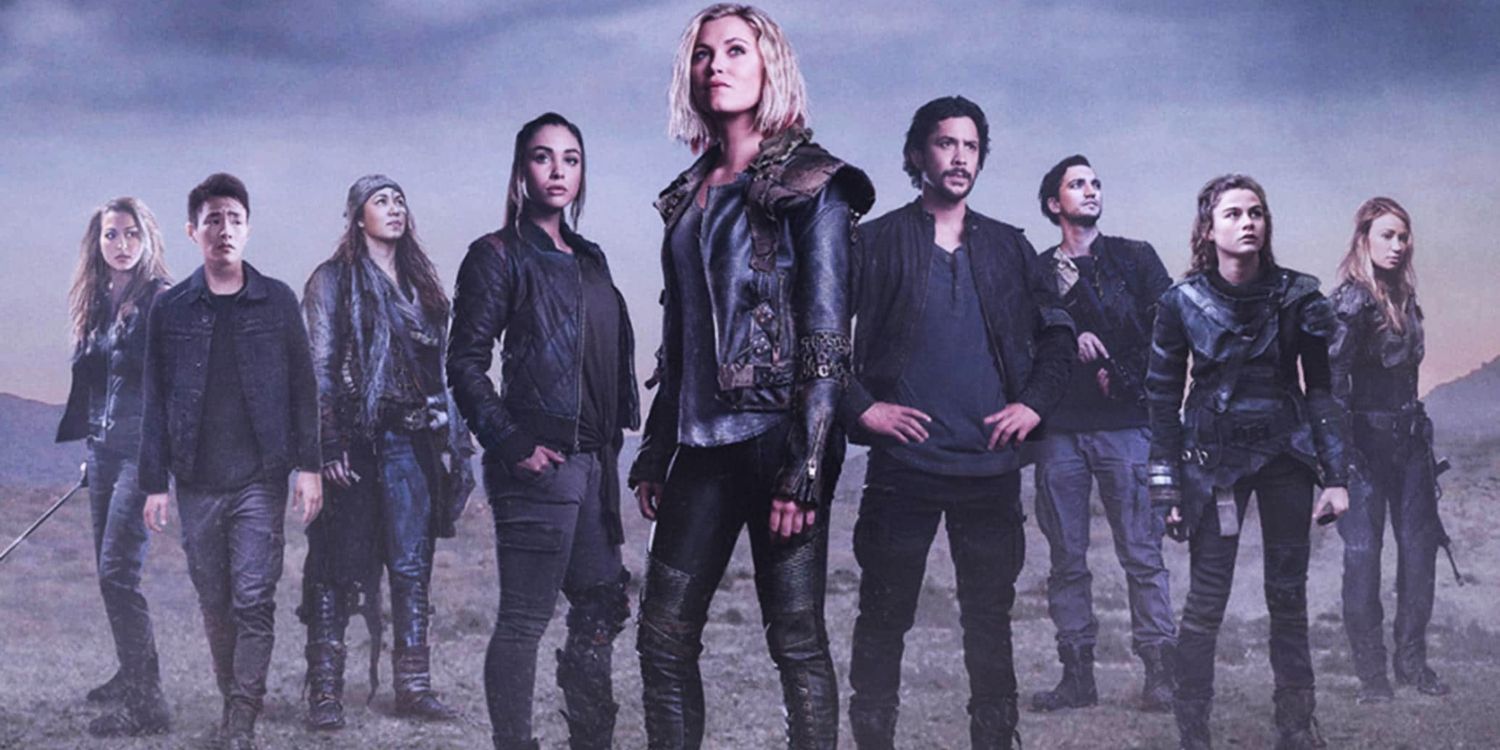 The 100 cast standing on dead grass wearing shades of grey and black for a season 5 promo shoot