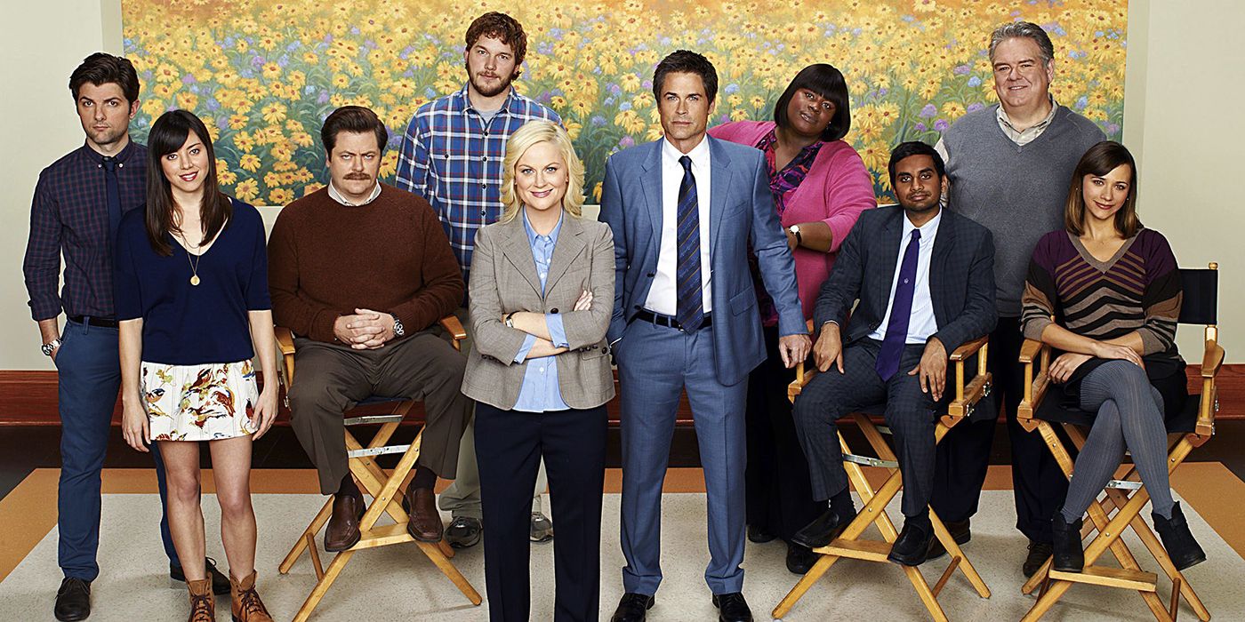 The cast of Parks and Recreation posing for a cast photo in front of a painting of flowers