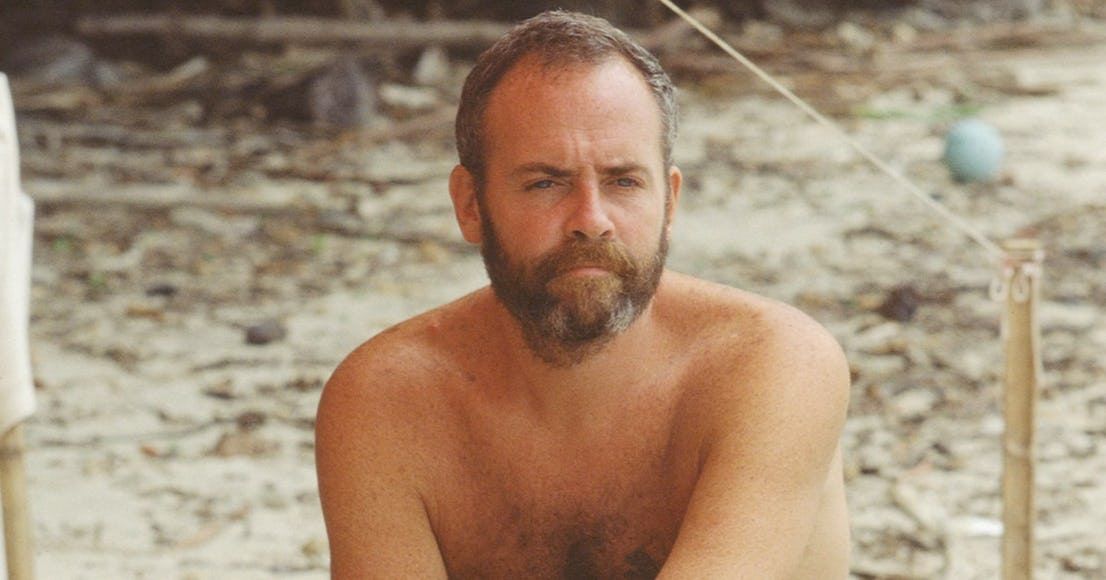 Richard Hatch sits at his tribe's beach with a concerned look on Survivor.