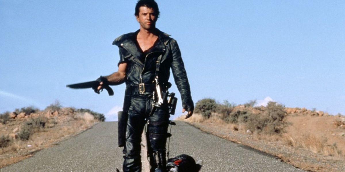 Max walks with his gun cocked as his dog lies on the road in The Road Warrior.