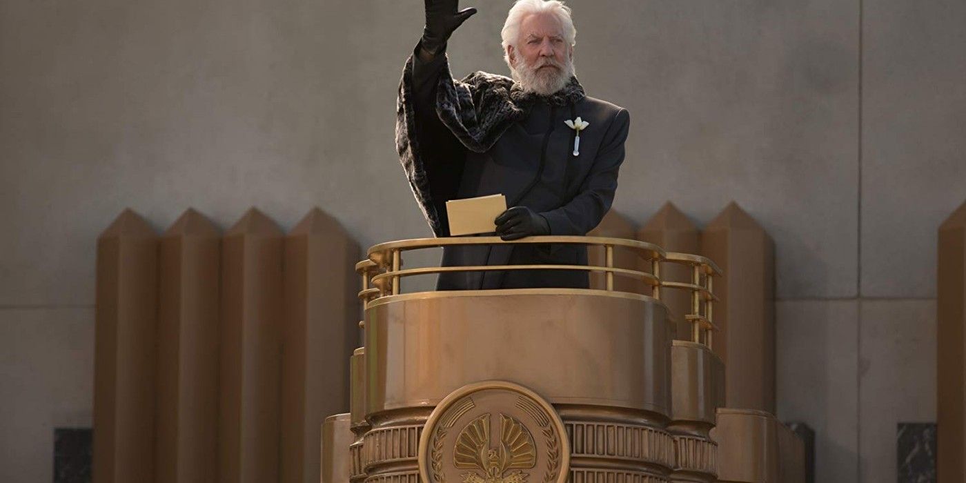 President Snow in black standing on a podium and raising his hand in the Hunger Games franchise.
