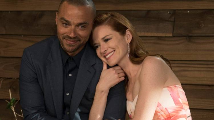 Jackson (Jesse Williams) and April (Sarah Drew) from Grey's Anatomy. The two smile against a wooden wall while April hugs Jackson by the arm.