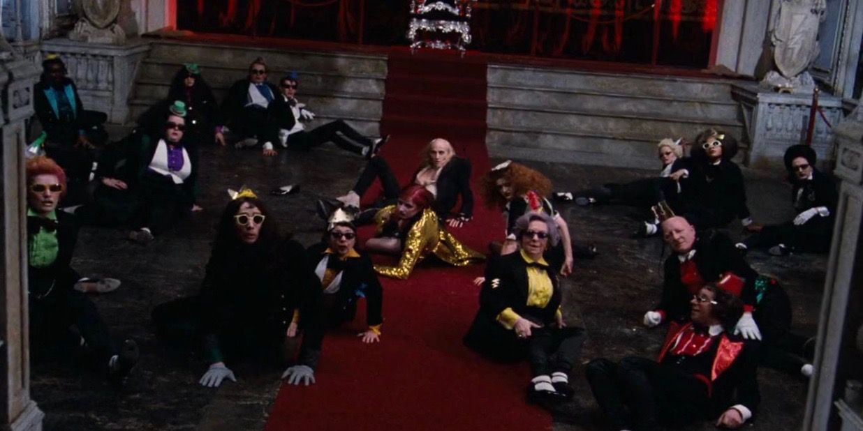 The cast of The Time Warp in The Rocky Horror Picture Show sitting and posing on a red carpet in front of a throne