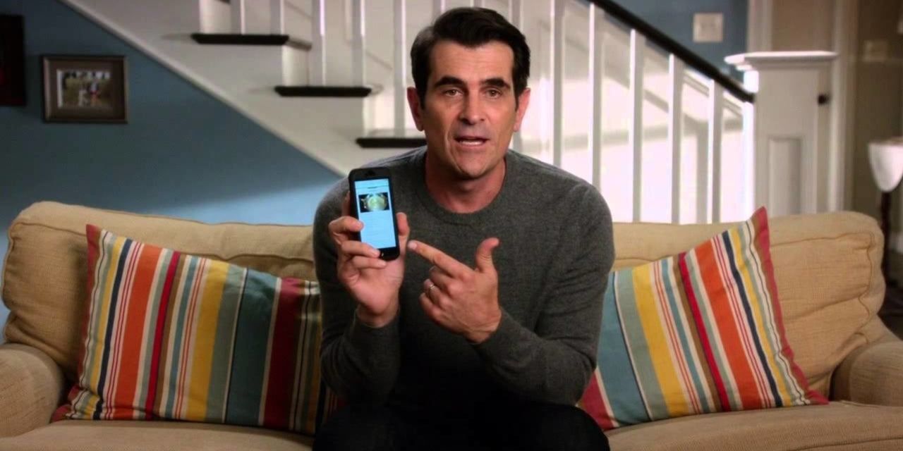 Phil sitting on the couch pointing to his phone on Modern Family