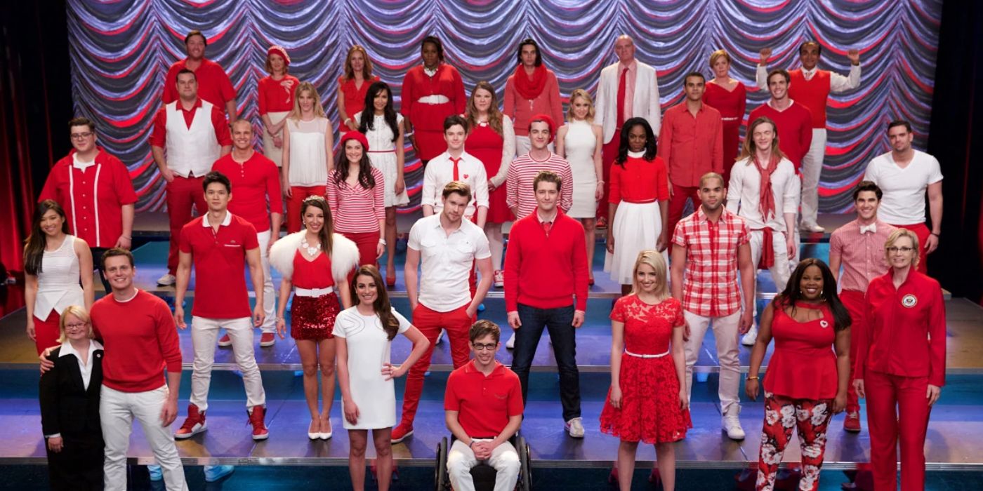 All the characters of Glee stand on the school stage, smiling at the camera