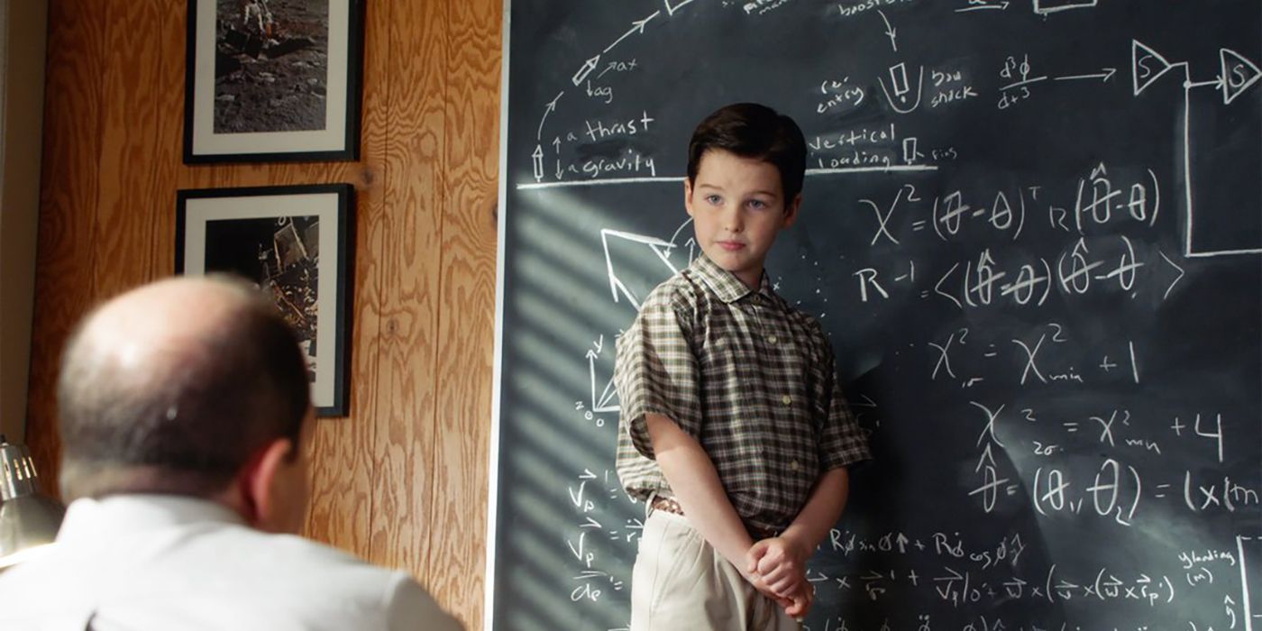 O jovem Sheldon na sala de aula ao lado do quadro-negro conversando com o professor.