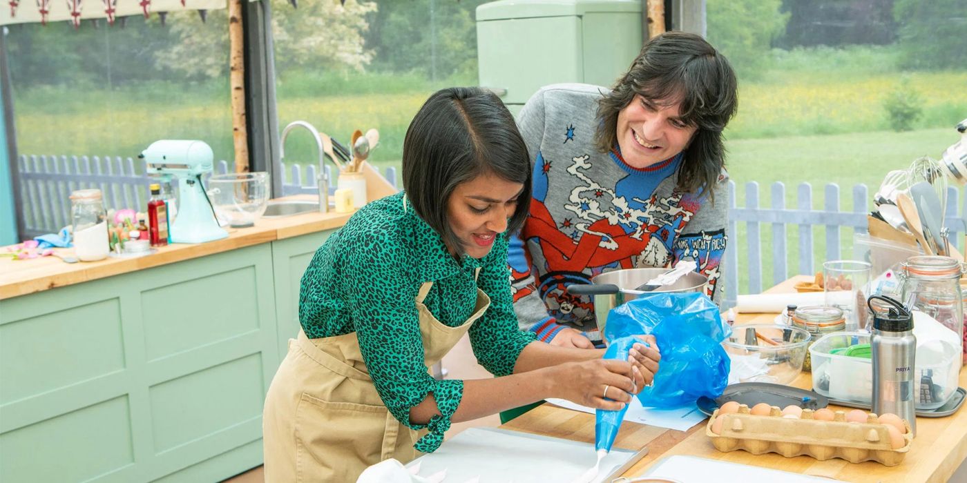 Noah Fielding in a grey printed sweater looking on as contestant Priya in a green top does the icing on a cake