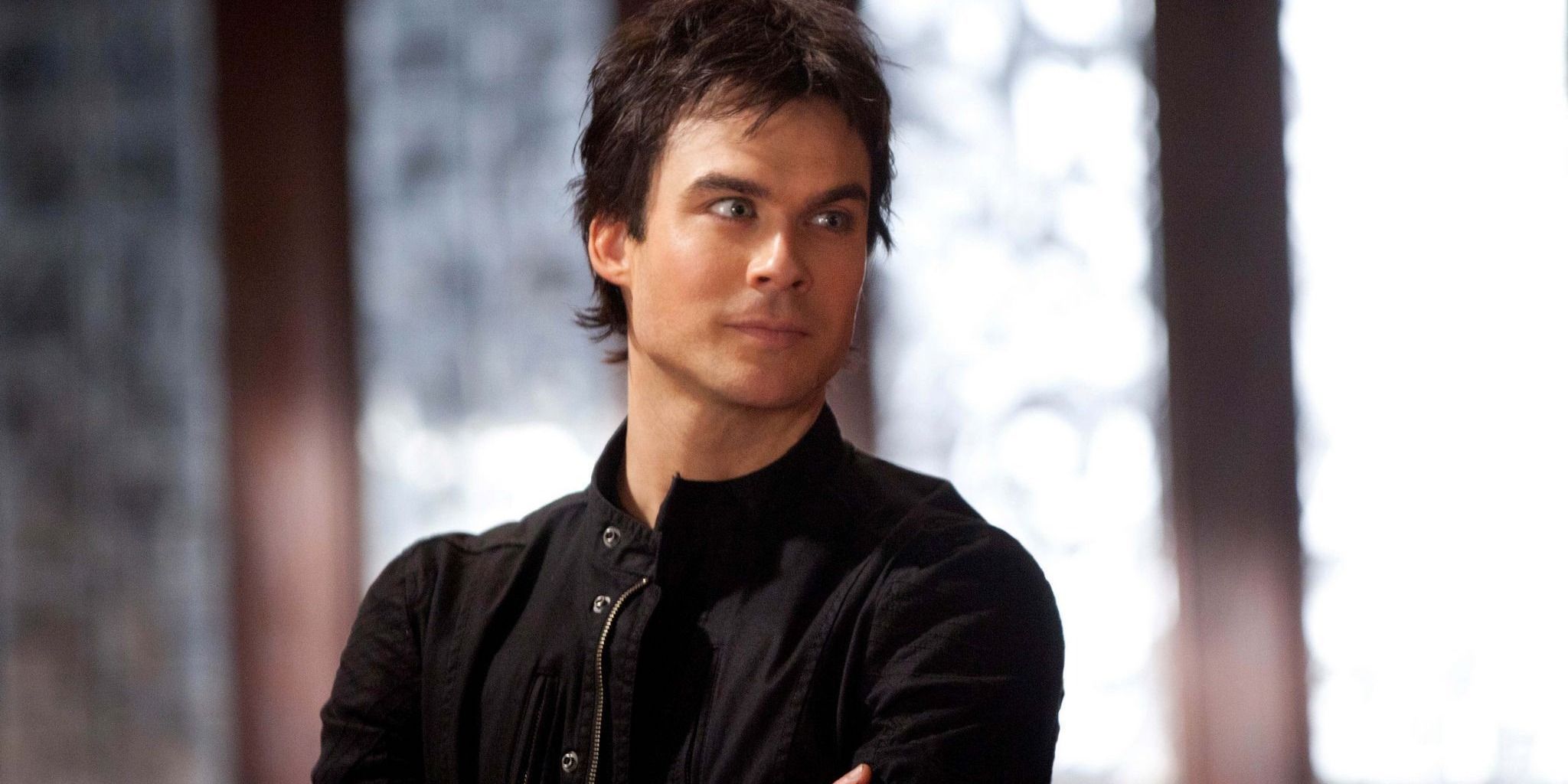 Damon standing with his arms crossed, looking at something with an amused expression.