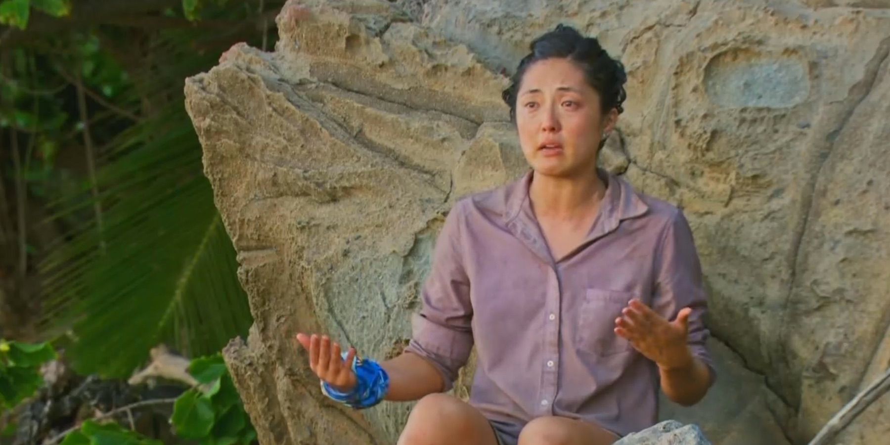Kellee Kim on Survivor, sitting on a rock wearing a sweater.