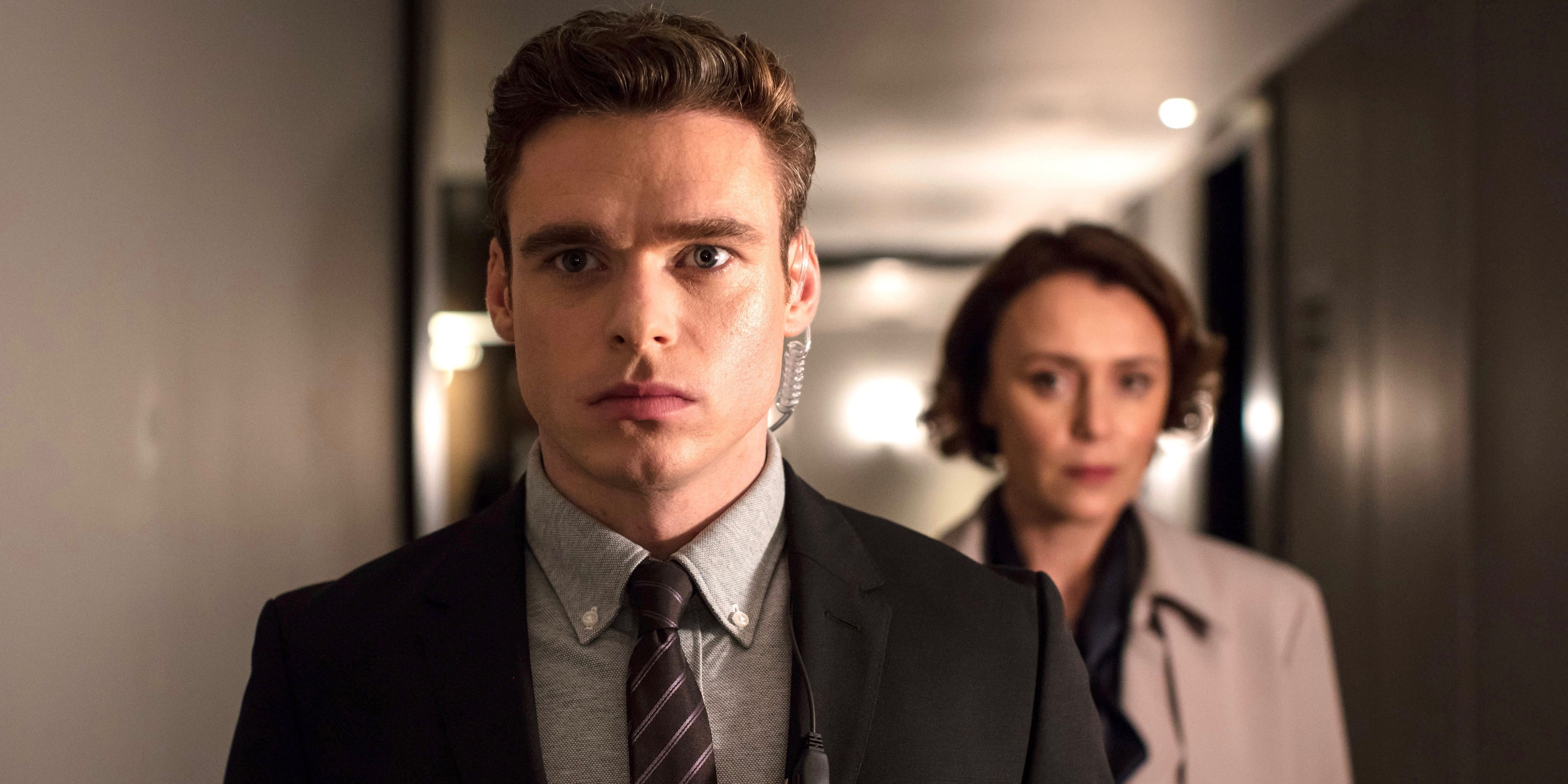 Richard Madden in Bodyguard with steely expression wearing earpiece with Keeley Hawes in background