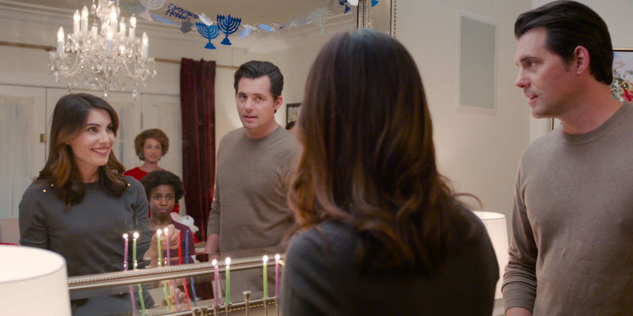 Rebeca And Chris stand in front of a mirror while lighting a menorah in Double Holiday