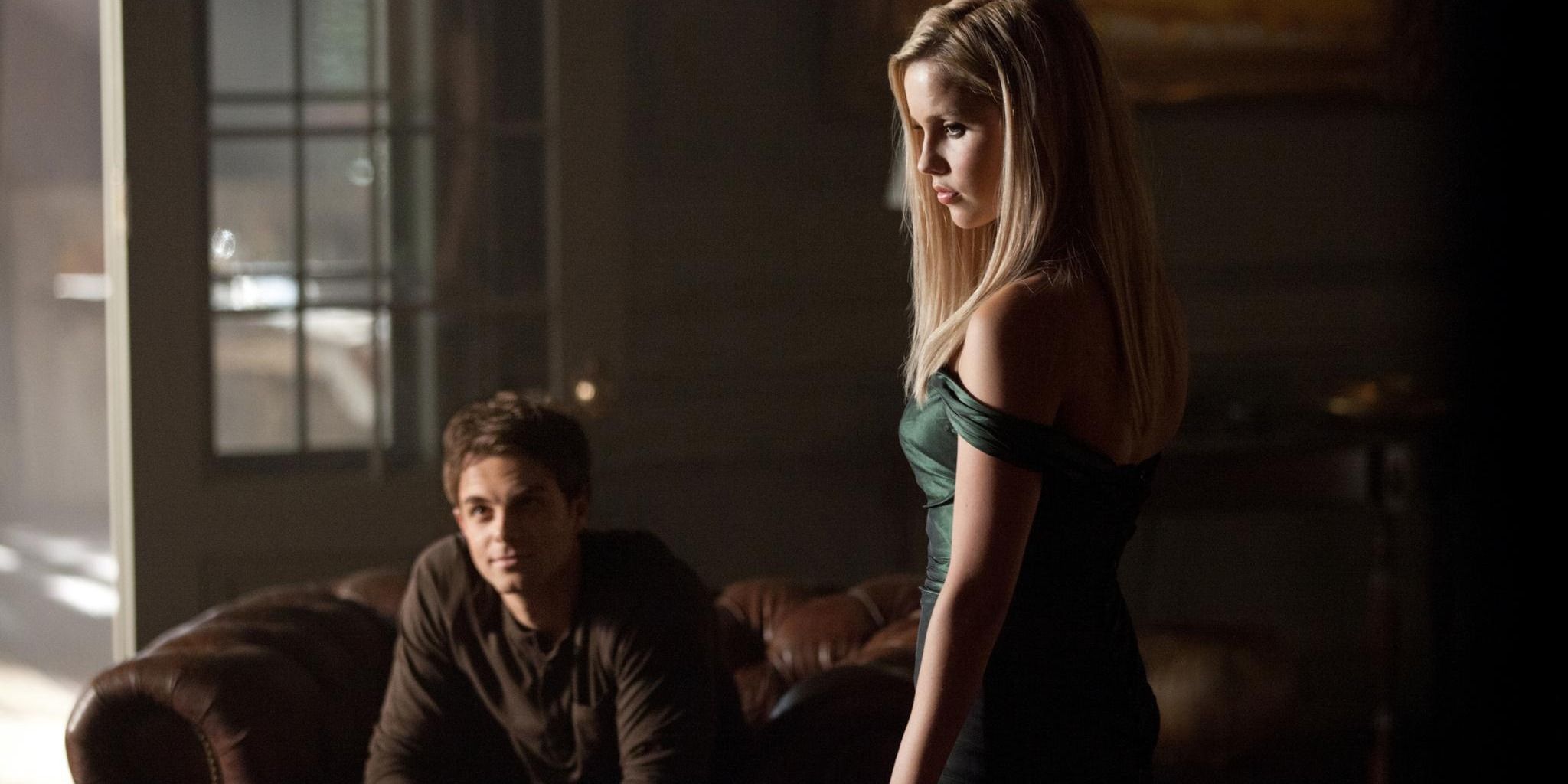 Rebekah em pé enquanto Kol Mikaelson se senta no sofá
