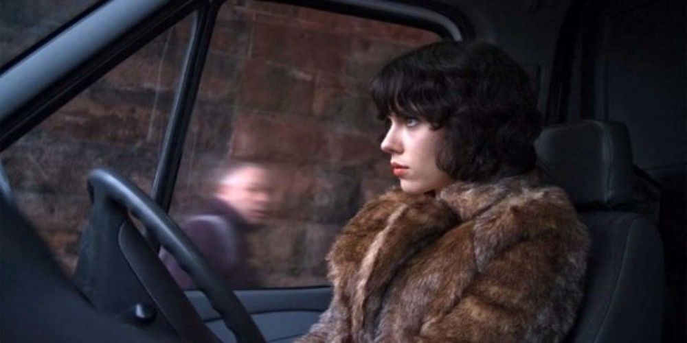 A woman in a fur coat sits in a car and stares straight ahead.