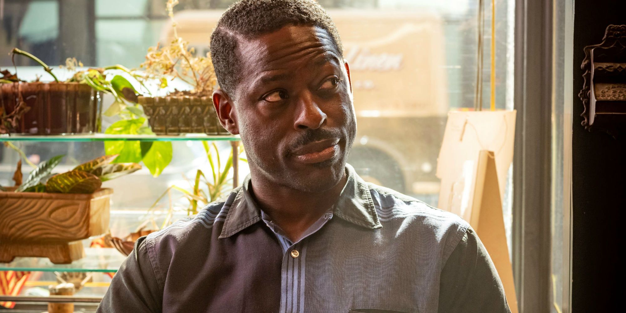 An image of Sterling K Brown in The Marvelous Mrs Maisel. He is seen to be sitting in a cafe and smiling to someone off-screen. He is also wearing a grey shirt