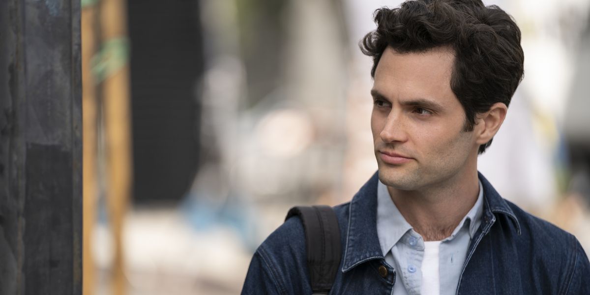 Joe standing to the side, wearing a jean jacket and looking off to the side smiling in a scene from You.