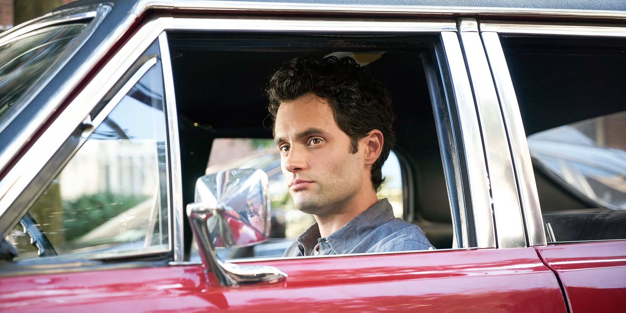 Joe sitting in a red car in a scene from You, looking out the window.