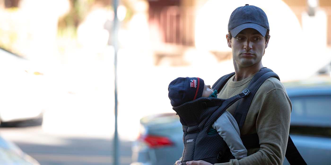 Joe wearing a baseball hat, carrying baby Henry in a front carrier in a scene from YOU.