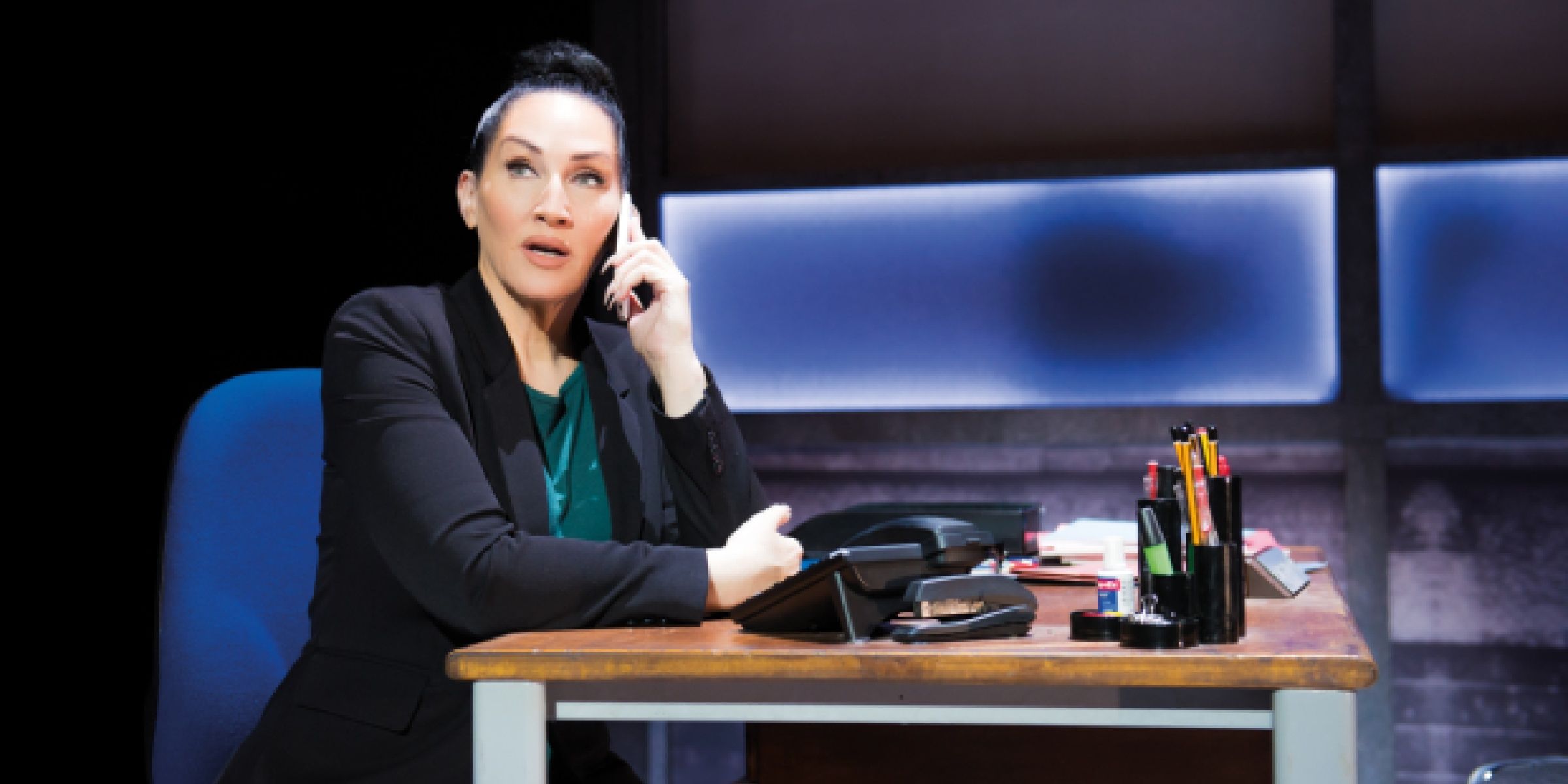 Michelle Visage as Miss Hedge sits behind a desk in the West End production of Everybody’s Talking About Jamie