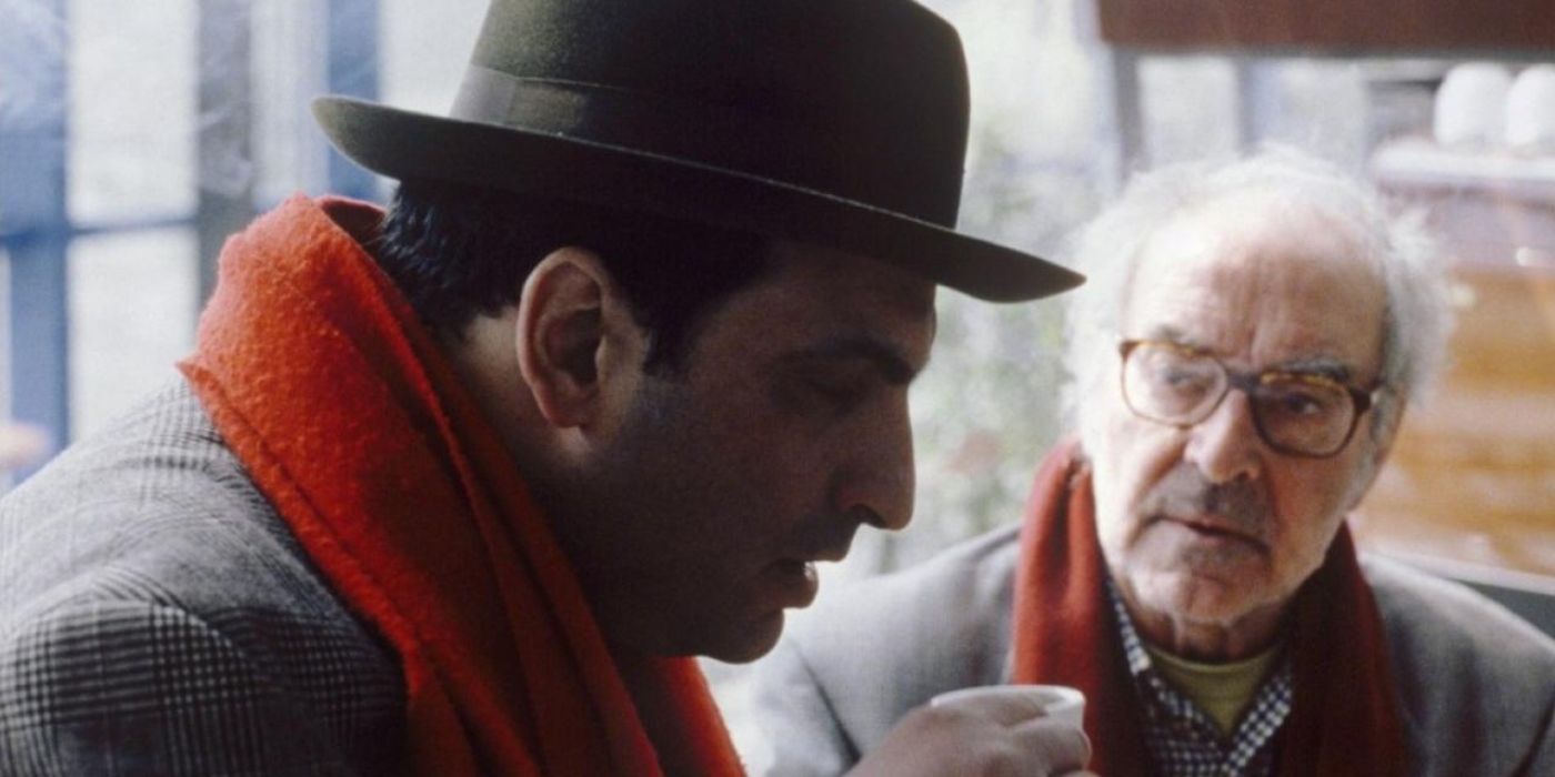 A man wears a hat and has tea as an older man watches in Notre Musique (2004).