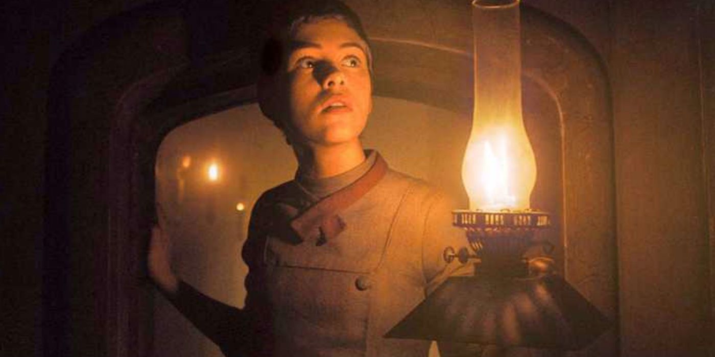 A young girl walks through a dark corridor. She is holding a lit oil lamp.