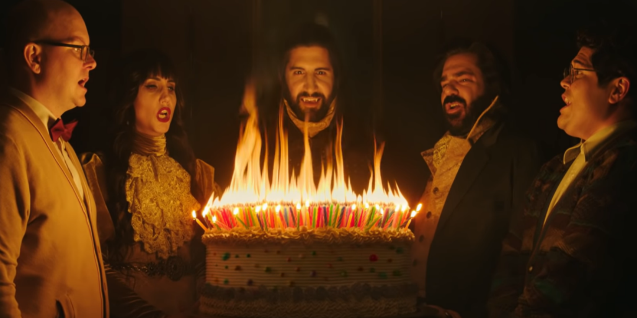 The cast members of What We Do In The Shadows standing around a birthday cake
