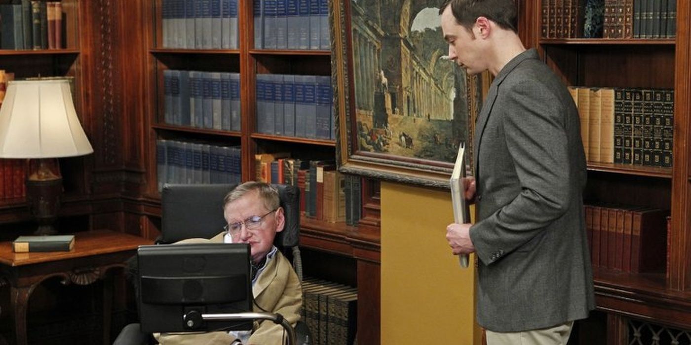 Stephen Hawking on his laptop in his wheelchair and Sheldon standing beside him in a grey suit on big bang theory