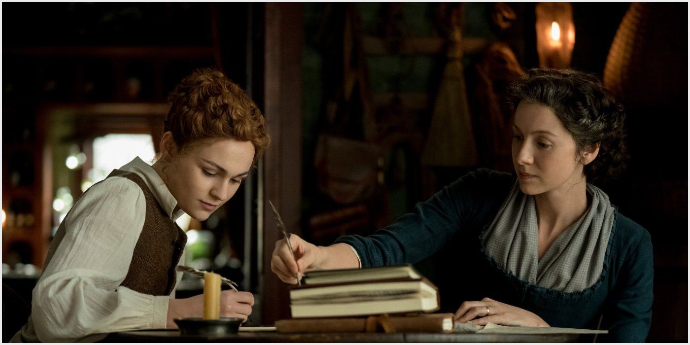 Brianna and Claire sitting together at a desk writing with quills in Outlander.