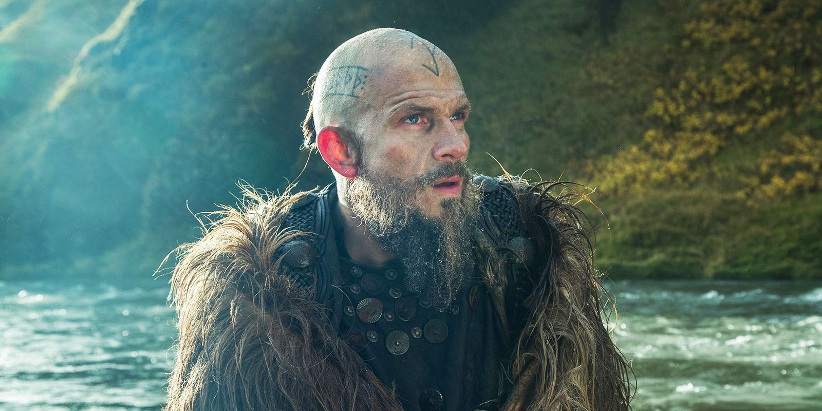 Floki in season 5's Vikings, standing in fur coat next to river.