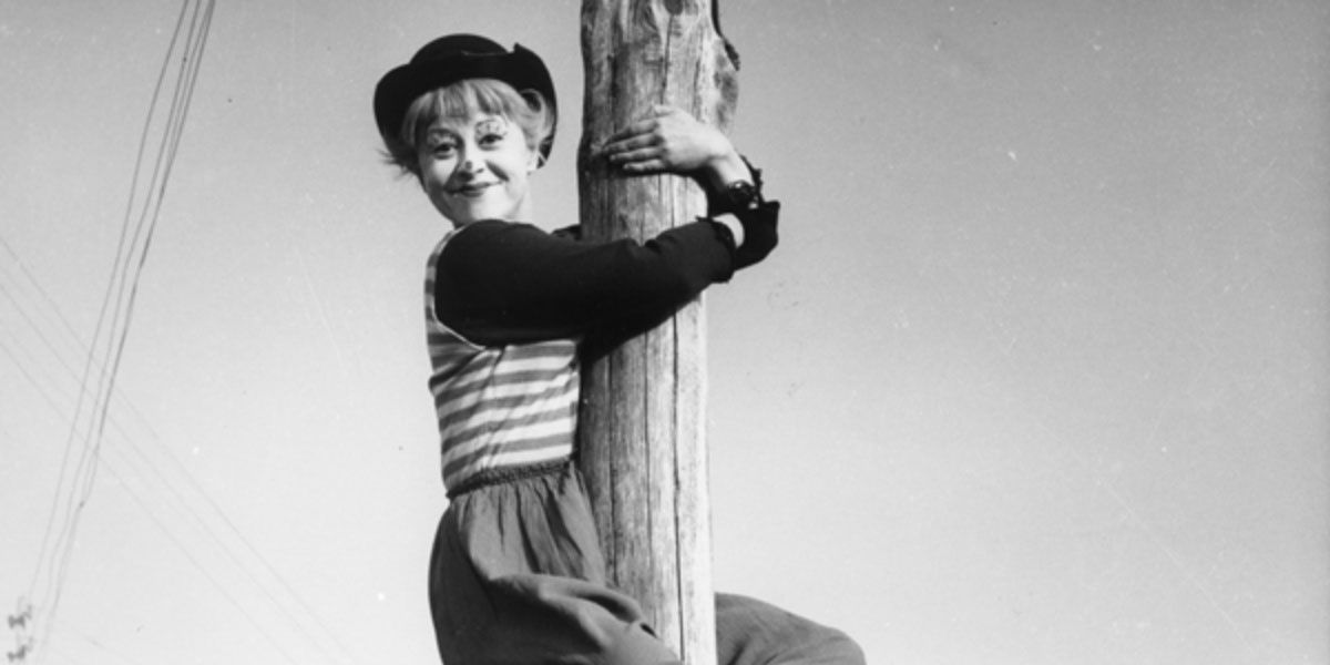 A woman in clown makeup climbs a post from La Strada