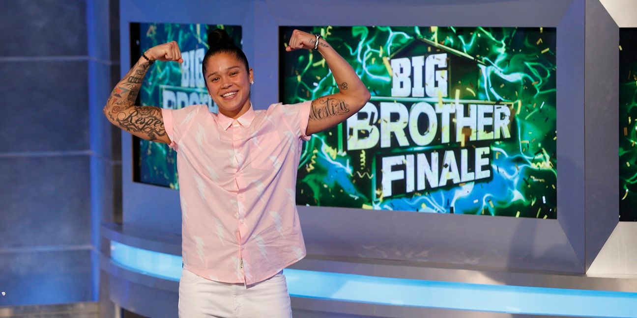 Kaycee flexionando seus músculos no final do Big Brother com uma tela atrás dela mostrando "Big Brother Finale" e o logotipo.  Ela está vestindo calças brancas e uma camisa rosa de manga curta.