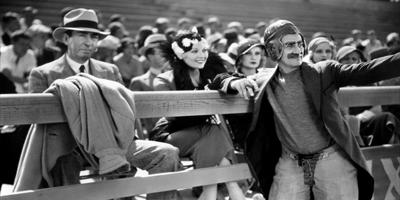 Groucho Marx em equipamento de futebol apoiado em uma cerca enquanto as pessoas olham em penas de cavalo