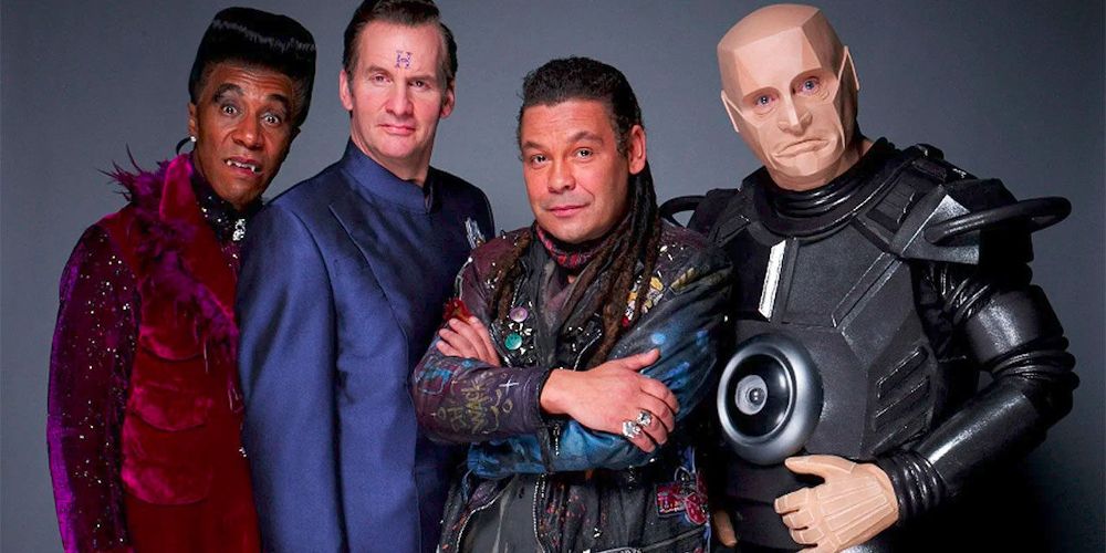The cast of Red Dwarf stand in front of a grey background, looking ready for action.