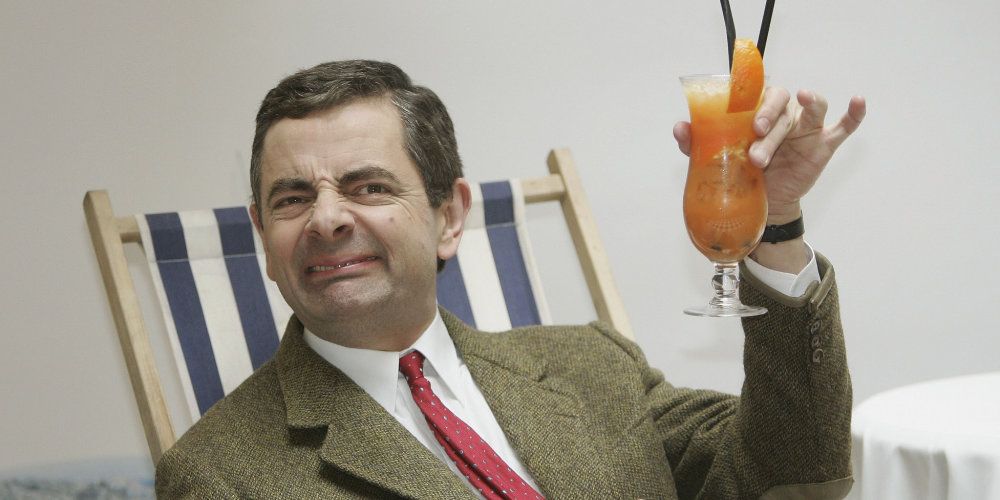 An image of Mr. Bean holding a drink while sitting in a seat 