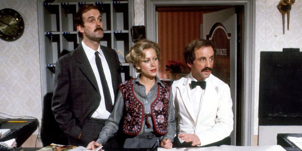 Basil Fawlty standing at the main desk with his colleagues in Fawlty Towers