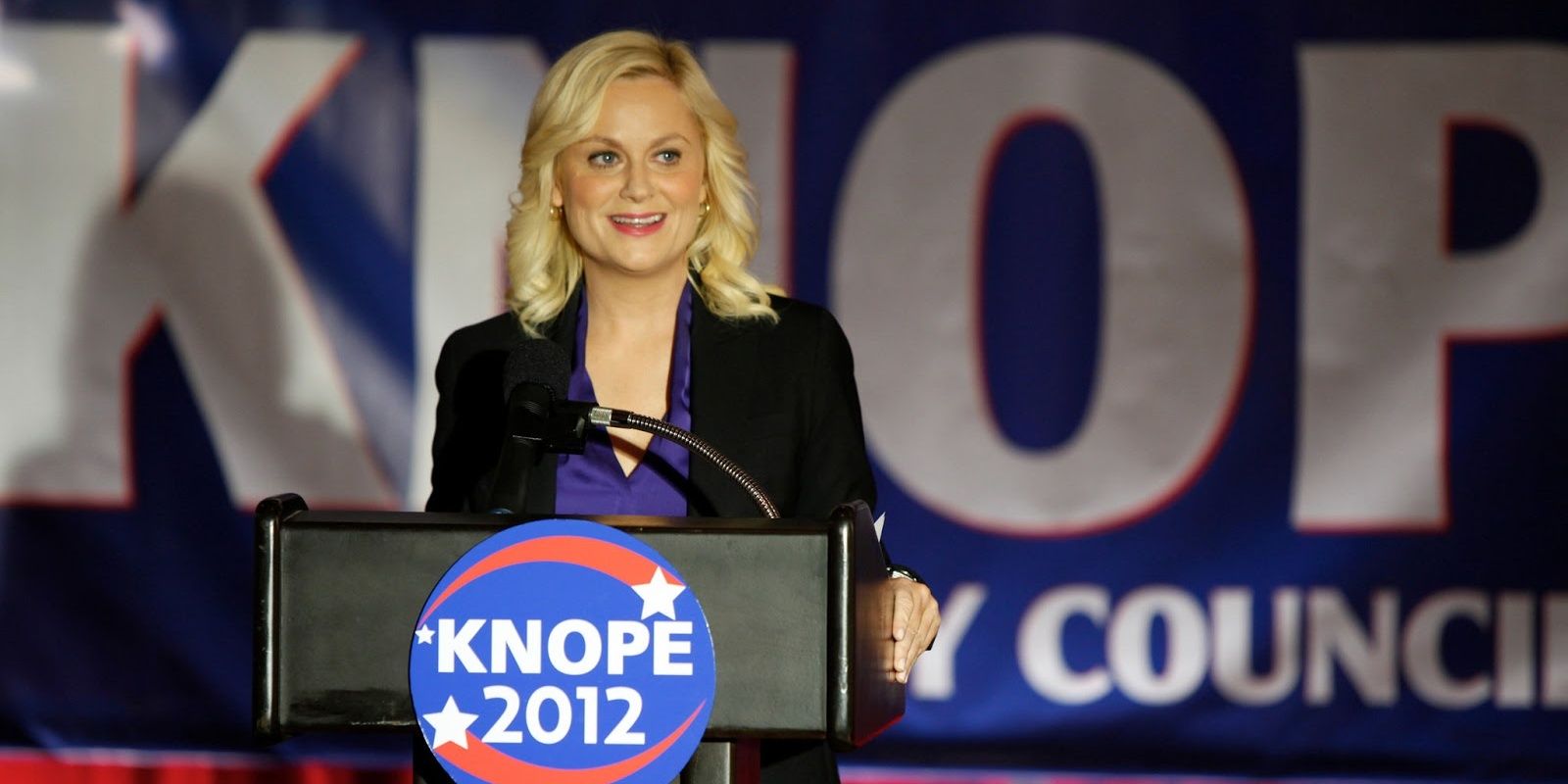 Leslie Knope standing at a podium in Parks and Recreation
