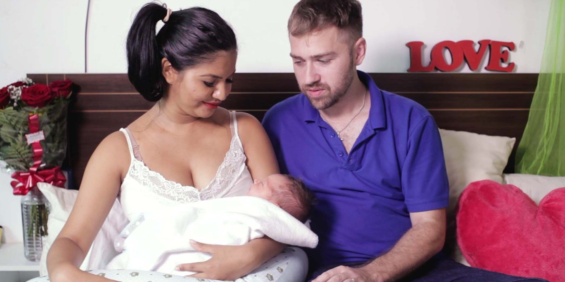 Paul Staehle and Karine Martins from 90 Day Fiance with their newborn son. Karine holds the baby as Paul looks on lovingly while seated on a bed