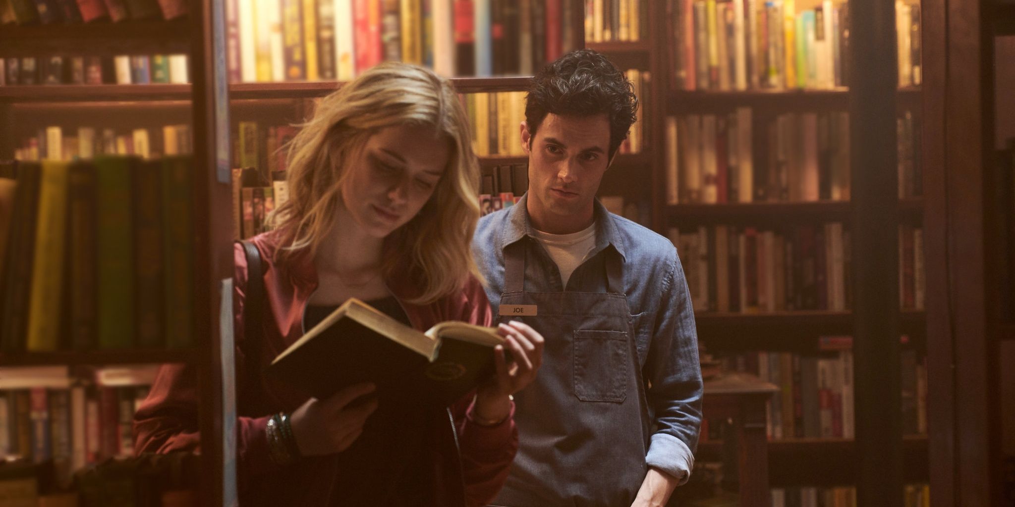 Beck reading a book while Penn Badgley's Joe stares at her from behind in You