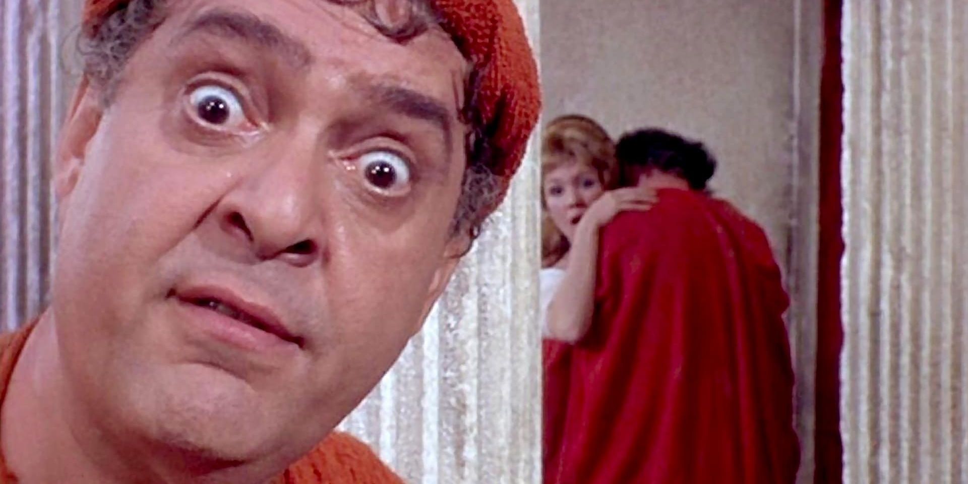 A character looking directly into the camera in A Funny Thing Happened On The Way To The Forum with two other characters embracing in the background
