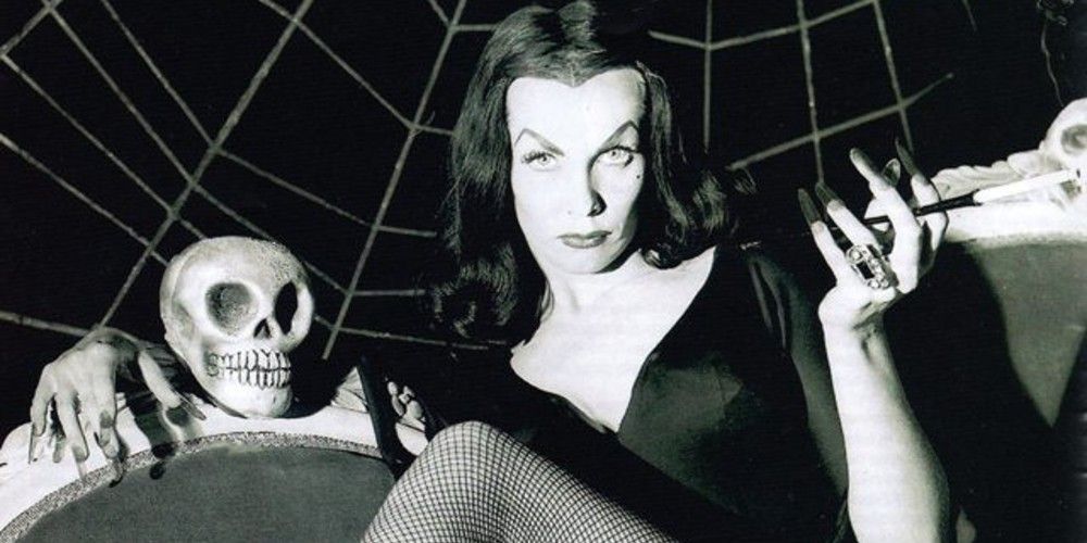 Vampira lounges on a couch with a cigarette next to a skull