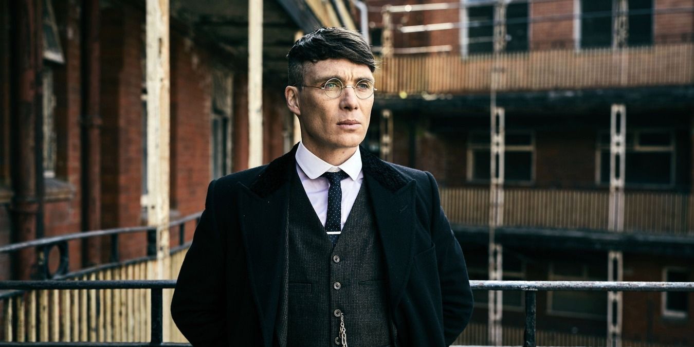 Thomas Shelby from Peaky Blinders in his suit and glasses standing on the balcony of a rundown building