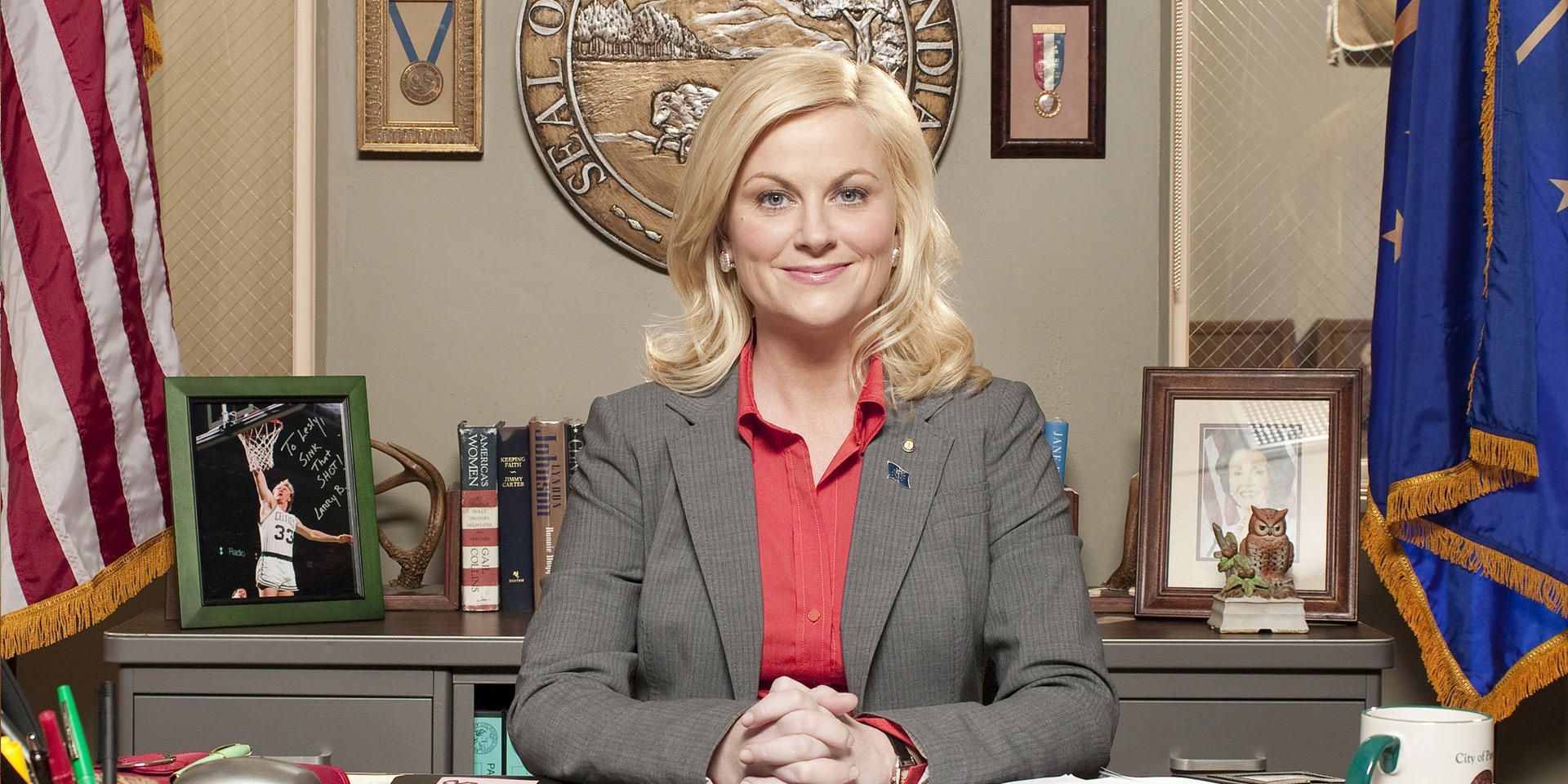Leslie Knope smiling and sitting behind her desk In Parks And Recreation