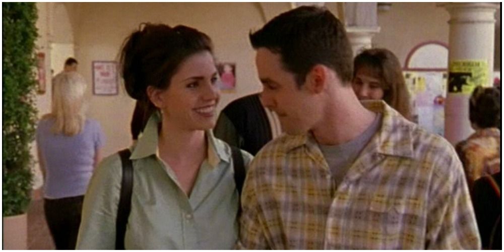 An image of Xander and Cordelia together while they are at school. Cordelia is seen wearing a green shirt and has her hair in a high pony. Xander is wearing a yellow and brown plaid shirt, with a grey t-shirt underneath. They are smiling at each other